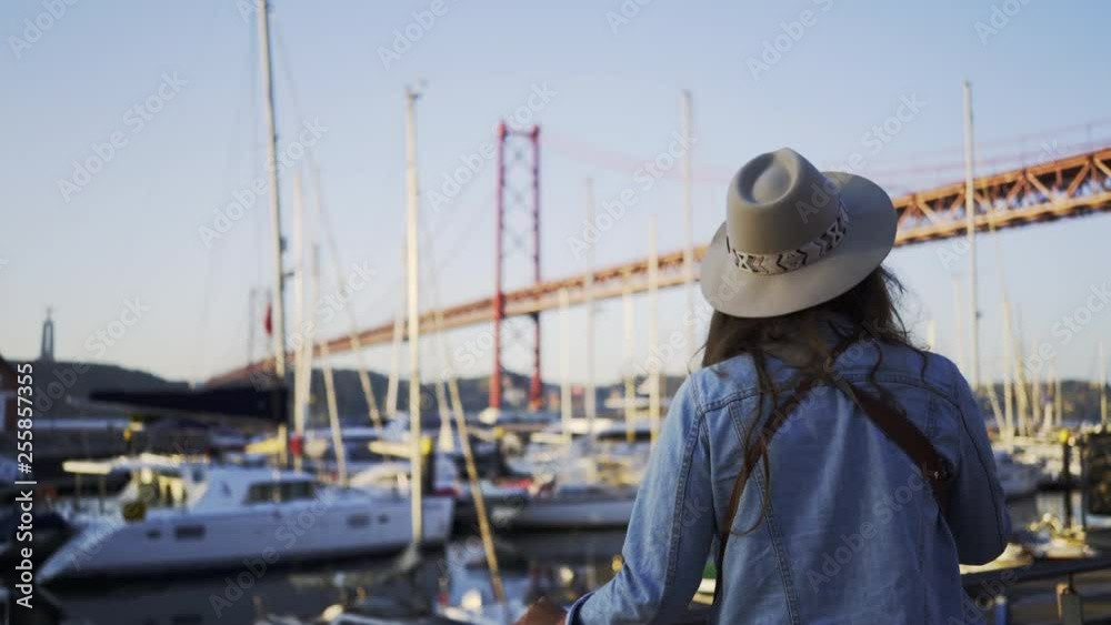 Back view woman browsing on smartphone while standing on embankment at background yachts in port and bridge 25th aplril. Lisbon, Portugal Back view woman browsing on smartphone while standing on embankment at background yachts in port and bridge 25th aplril. Lisbon, Portugal