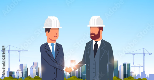 two businessmen builders handshake agreement during meeting over city construction site tower cranes building residential buildings cityscape sunset skyline background portrait horizontal