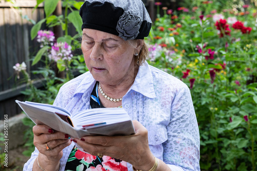 Older Jewish woman wearing jewelry reads her prayer book in backyard garden. photo