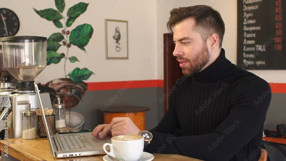 Successful serious man in a black sweater working on a laptop with documents in a coffee shop during lunch, next to the computer is a cup of latte with a pattern. Successful serious man in a black sweater working on a laptop with documents in a coffee shop during lunch, next to the computer is a cup of latte with a pattern.