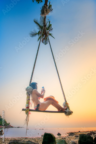 Young woman enjoy and happiness on sway the wooden swing under coconut palm tree over the sea beach, sunbeam and sunset light relaxation