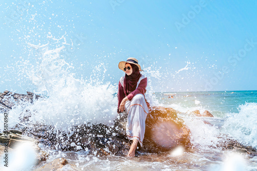 confident young muslim asian woman sitting on rock with beautiful wave splash background . woman sitting on cliff over sea. Travel concept.