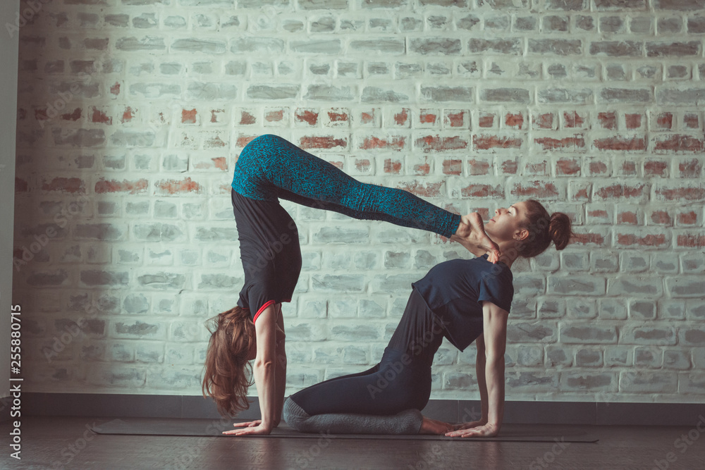 two beautiful caucasian women doing partner yoga in yoga studio opposite brick wall