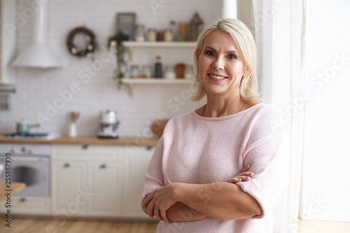 Attractive middle aged housewife with blonde hair and brown eyes posing indoors in her modern clean stylish kitchen interior, crossing arms on her chest and looking at camera with pleased smile photo