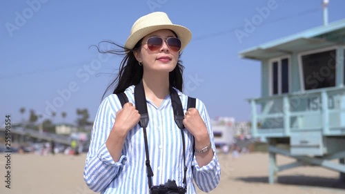 young asian girl photographer carrying professional slr camera traveling california usa. woman in sunglasses standing on beach sand sightseeing sea view. american flag waving on lifeguard stand