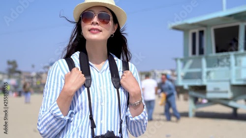 young korean girl traveler carrying backpack standing on sandy beach relaxing enjoy sunny day on summer vacation lifestyle. people walking in background near lifeguard tower Santa Monica Pier.
