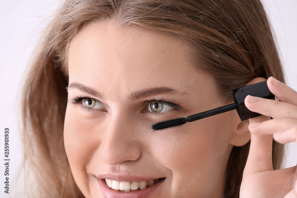 Young woman applying mascara onto eyelashes against light background, closeup