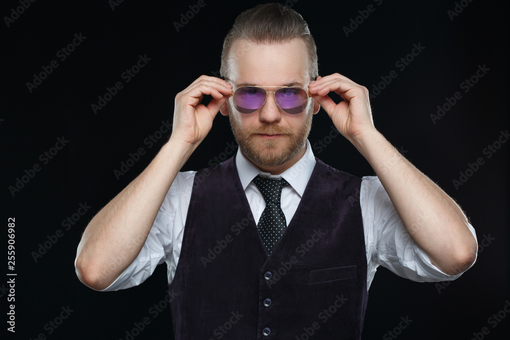 Handsome man with a beard straightens his sunglasses sunglasses isolated on a black background.