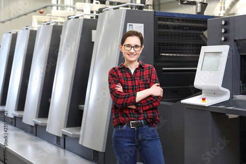 Praca w drukarni. Uśmiechnięta kobieta, pracownik drukarni stoi w hali produkcyjnej na tle nowoczesnych maszyn drukarskich