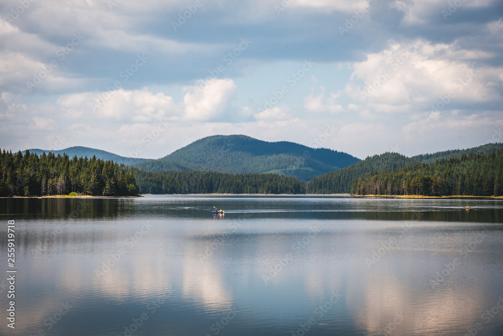 Fototapeta premium Beautiful summer landscape - calm waters of a lake and a boat. Green forest and mountain in the background. Bulgaria.