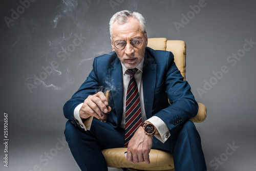 pensive mature businessman in suit and glasses holding cigar while sitting in armchair on grey