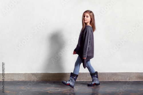 Fashion kid casual stylish clothes. Model child walks, pose. little girl poses near white wall, wears leggings, sweater, rubber boots. Full length portrait . Copy space.