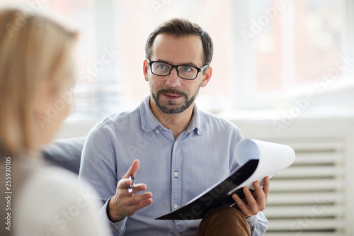 Young confident counselor with pen and document explaining something to his patient during conversation
