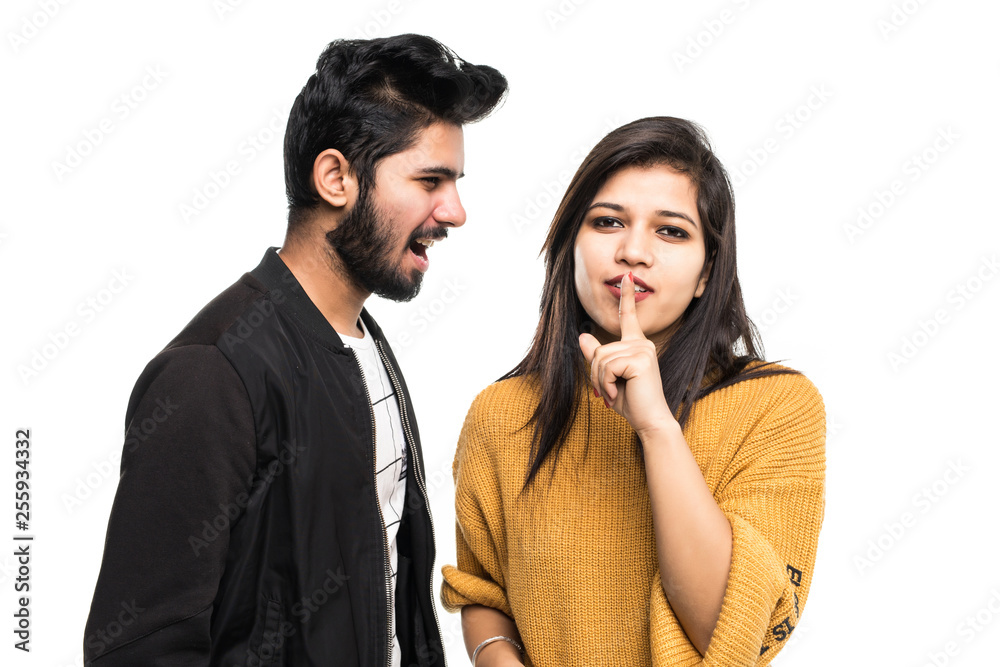 Portrait of a smiling indian couple, man quarrel but woman showing silence gesture and looking at camera over gray wall Portrait of a smiling indian couple, man quarrel but woman showing silence gesture and looking at camera over gray wall