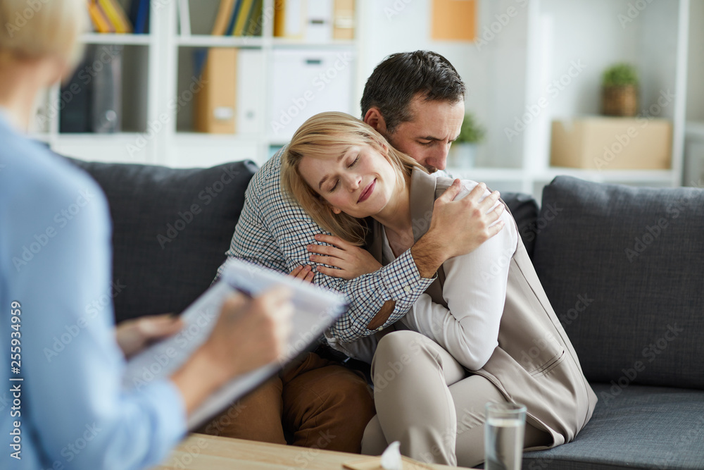 Young couple sitting on couch in embrace after consulting with counselor and solving their problem Young couple sitting on couch in embrace after consulting with counselor and solving their problem