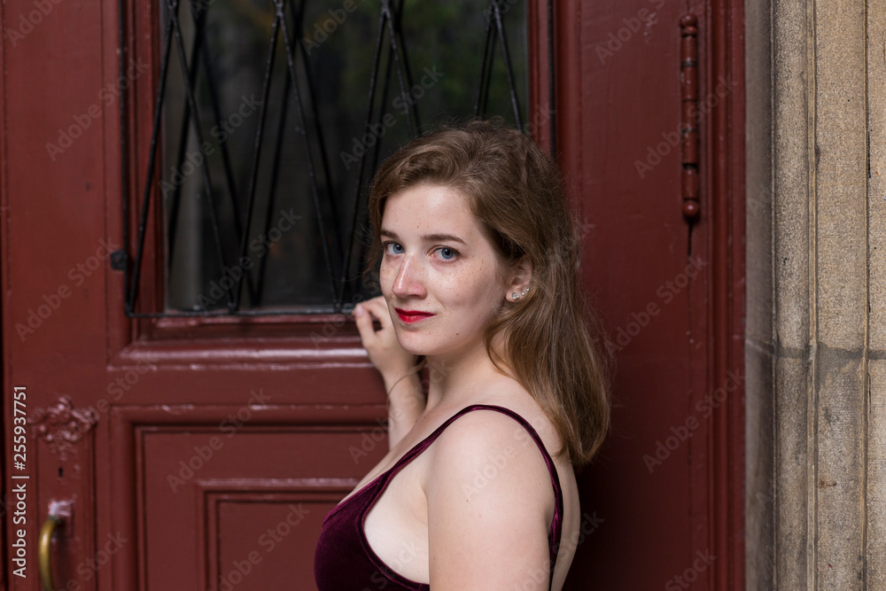 Fototapeta premium Medium horizontal portrait of exquisite blue-eyed young woman in velvet tank top with small smile turning while standing against dark red door