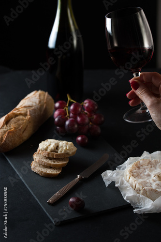 hand holding a glass of red wine,  a wine bottle, brie cheese, cluster  grapes, rustick bread with anrique knife. photo