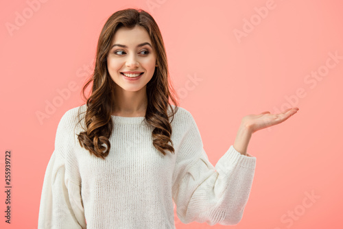 smiling and beautiful woman in white sweater pointing with hand isolated on pink