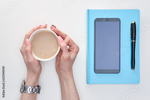 Woman's hands holding cup of latte, blue covered diery with ink pen and smartphote above, white background, view from top photo