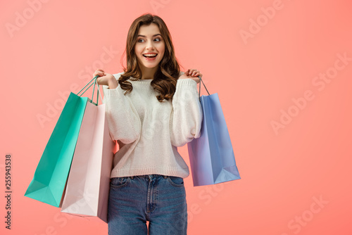 smiling and attractive woman in white sweater holding shopping bags isolated on pink