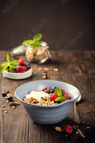 Oat granola with berries and yoghurt in blue bowl on dark old wooden background. Copy space. Healthy breakfast concept.
