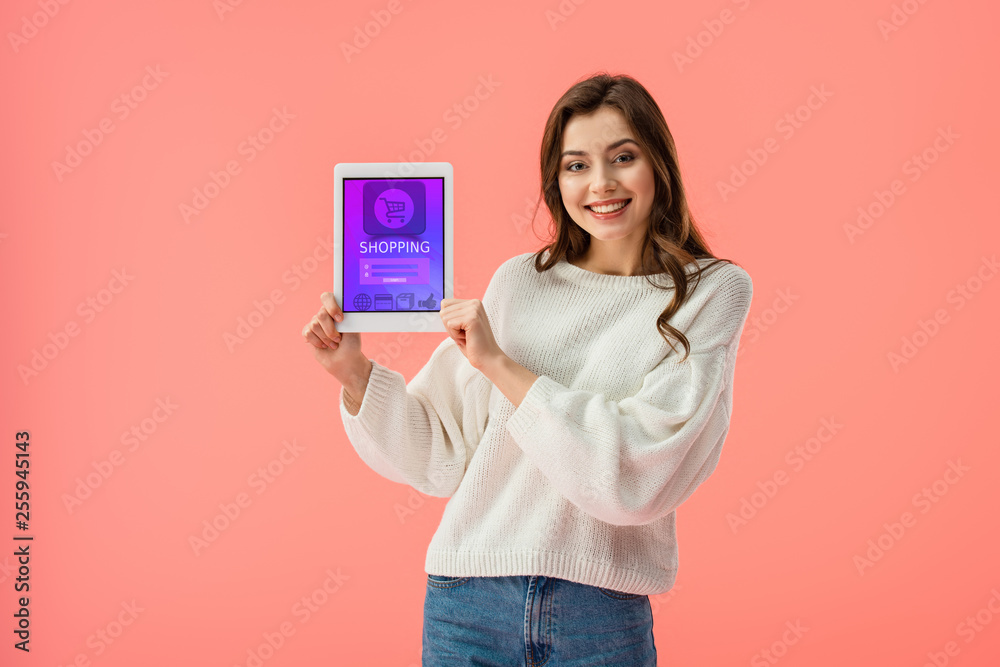 cheerful young woman holding digital tablet with shopping app on screen isolated on pink