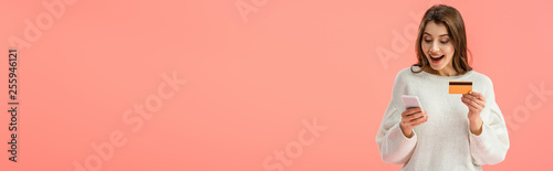 panoramic shot of surprised brunette girl holding credit card and smartphone isolated on pink