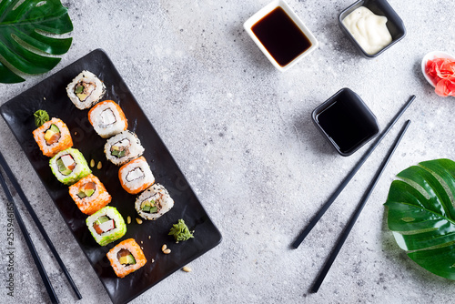 Various Fresh and delicious sushi set on black and white slate with metal and slate sticks, sauce and green leaves on light stone background , flat lay