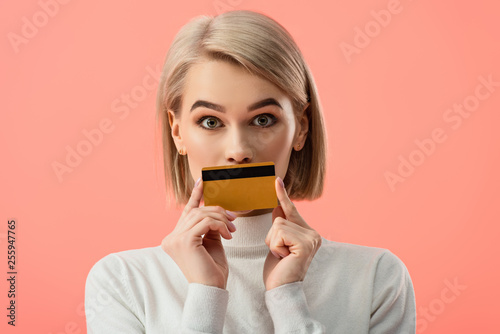 surprised blonde woman covering mouth with credit card isolated on pink photo