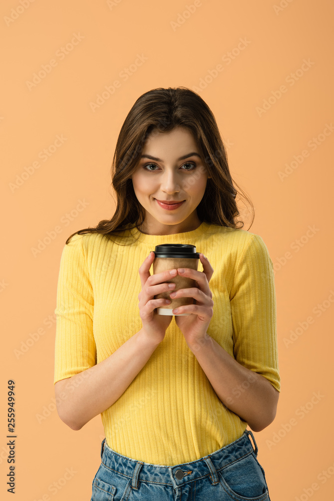 Front view of tricky brunette girl holding paper cup of coffee isolated on orange Front view of tricky brunette girl holding paper cup of coffee isolated on orange