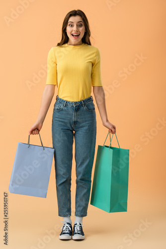 Full length view of surprised smiling brunette girl holding shopping bags on orange background