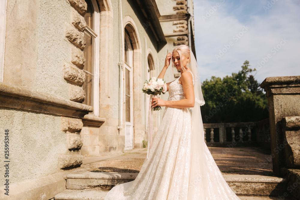 Obraz premium Gorgeous bride in wedding dress standing on the stairs by the palace. She hold a bouquet of flowers