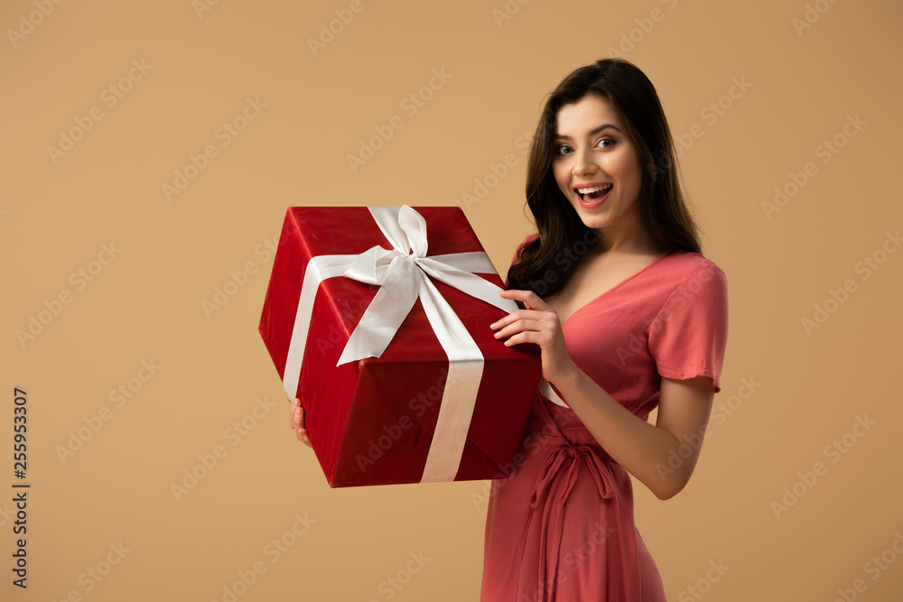 Surprised brunette girl in dress holding red gift box and smiling isolated on beige Surprised brunette girl in dress holding red gift box and smiling isolated on beige