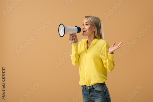 Smiling blonde woman in yellow shirt screaming in megaphone isolated on beige