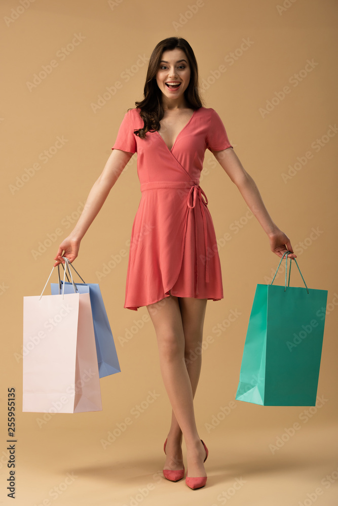 Laughing brunette girl in dress holding shopping bags and standing with crossed legs on beige background