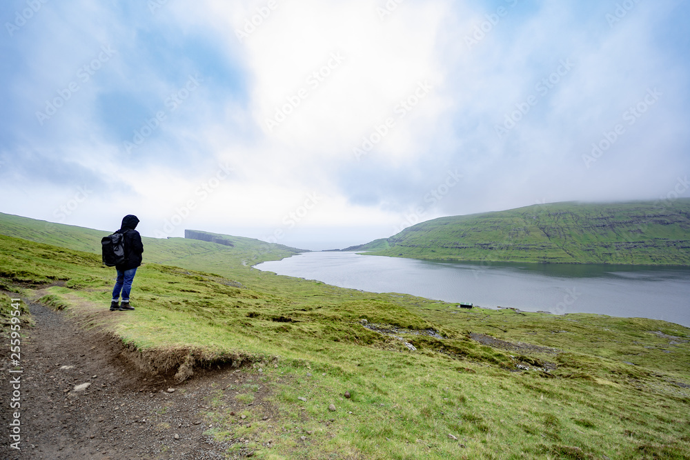 Young man standing while trekking on rural road beside the lake with cloudy weather raining in Faroe Islands, north Atlantic ocean, Europe