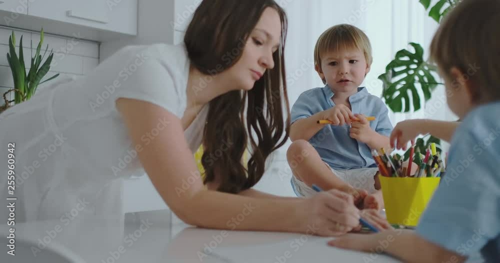 Mom helps sons learn to draw doing homework preschool preparation at home sitting in the white kitchen. Two children brothers draw a portrait of mother together Mom helps sons learn to draw doing homework preschool preparation at home sitting in the white kitchen. Two children brothers draw a portrait of mother together
