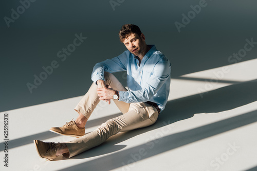 handsome man in blue shirt and beige jeans sitting in sunlight and looking at camera