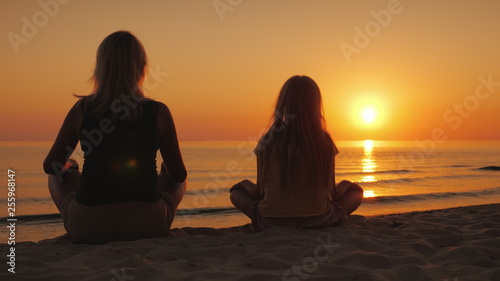 A woman with her daughter sitting side by side on the sand in a lotus pose, looking at the sunset over the sea