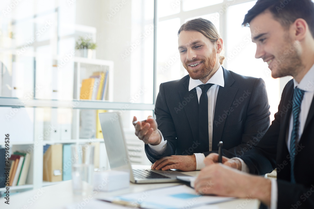 One of cheerful successful brokers pointing at laptop display while making presentation of new business strategies One of cheerful successful brokers pointing at laptop display while making presentation of new business strategies