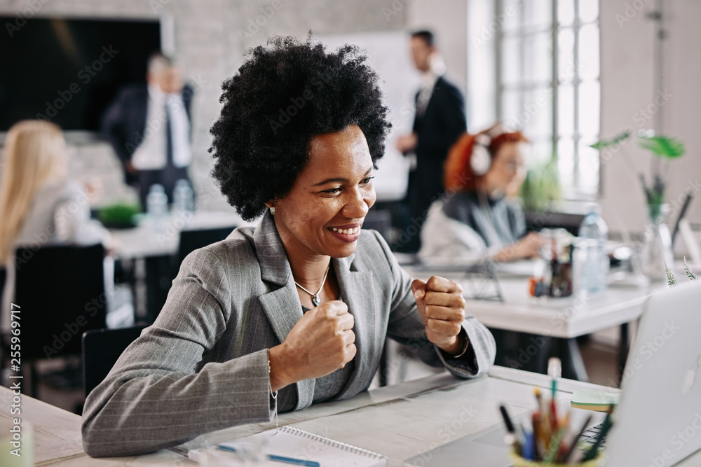 Happy African American businesswoman celebrating success while using laptop at her desk.