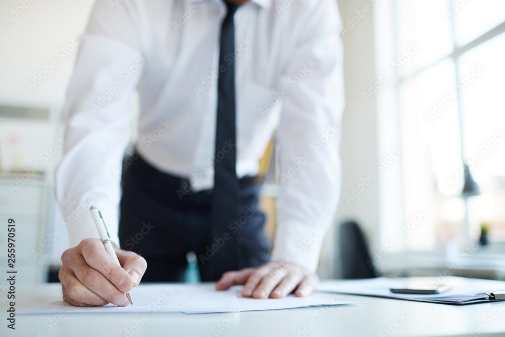 Young businessman with pen signing paper or contract while bending over desk in office after negotiation Young businessman with pen signing paper or contract while bending over desk in office after negotiation