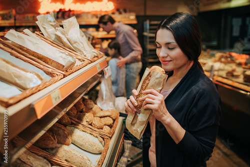 Young parents and daughter in grocery store. Cheerful woman smell bread she hold in hands. Father stand behind with daughter and carry grocery trolley.