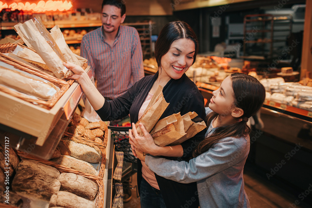 Young parents and daughter in grocery store. Cheerful mother and girl pick upb bread and rolls together. They smile to each other. Man stand behind with trolley.