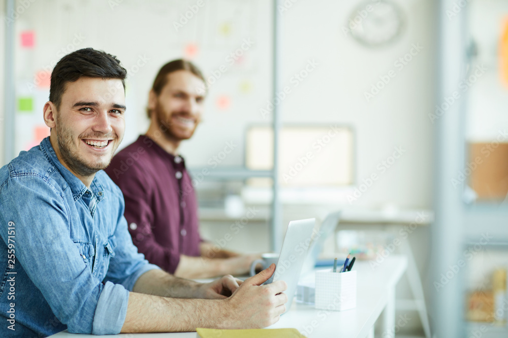 Cheerful young manager with toothy smile and his colleague in casualwear looking at you by workplace Cheerful young manager with toothy smile and his colleague in casualwear looking at you by workplace