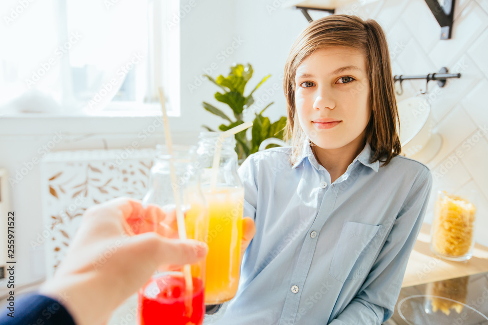 Smiling little preteen boy with long straight hair clinking glasses with fresh juice in sunny morning in kitchen. Smiling little preteen boy with long straight hair clinking glasses with fresh juice in sunny morning in kitchen.