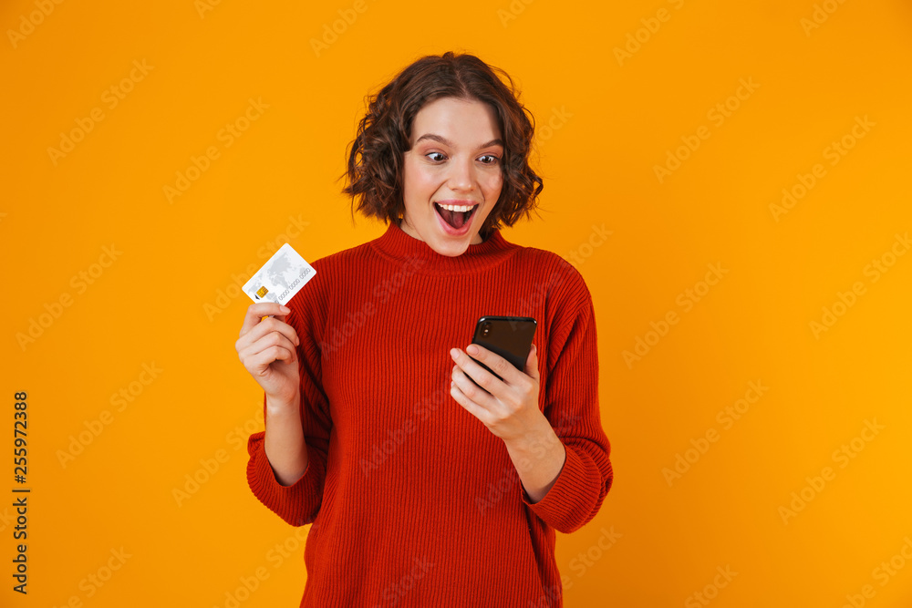 Portrait of joyous woman using cell phone and credit card while standing isolated over yellow background Portrait of joyous woman using cell phone and credit card while standing isolated over yellow background