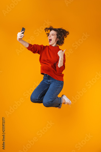 Portrait of positive woman jumping and taking selfie photo on cell phone while standing isolated over yellow background © Drobot Dean