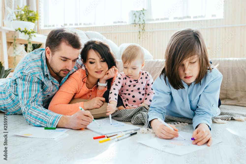 Charming family of four: father, mother, son, daughter drawing together, lying on the floor in a beroom. New home, happy healthy relationship concept.