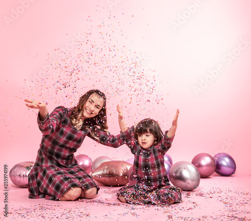 Funny mom and child sitting on a pink background. Little girl and mother having fun with balloons and confetti